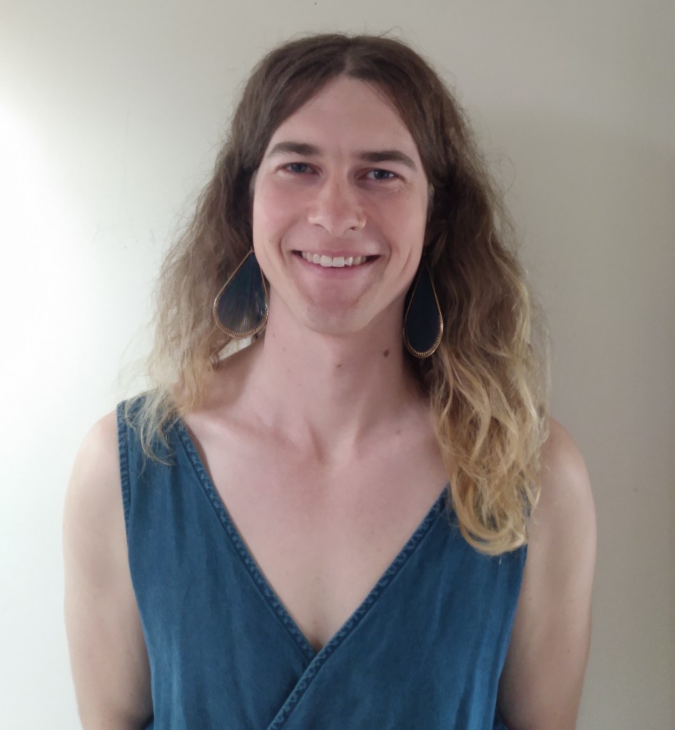 Photograph of Akki, a white transfemme and nonbinary person, smiling directly into the camera. They have long ombre blonde hair and are wearing hoop earrings and a blue v-neck top.