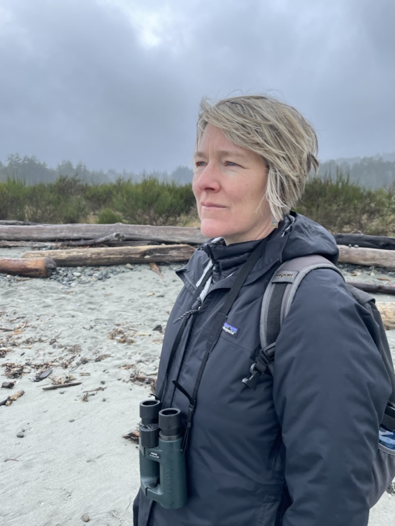 Photograph of Cindy, a white cisgender woman with short blonde hair, standing on a sandy beach while wearing a black jacket, a grey backpack, and binoculars around her neck. In the background of the photo, there is some driftwood and a cloudy sky. Cindy is looking pensively beyond the camera.
