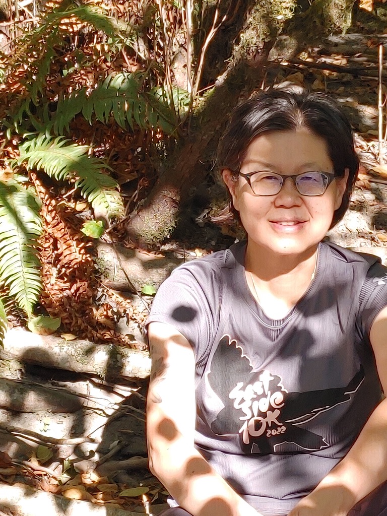 Photograph of Fionna, a Chinese cisgender woman with short black hair tucked behind her ears, sitting in dappled sunlight in a forest setting and smiling. Fionna is wearing a grey East Side 10K 2022 t-shirt and glasses.