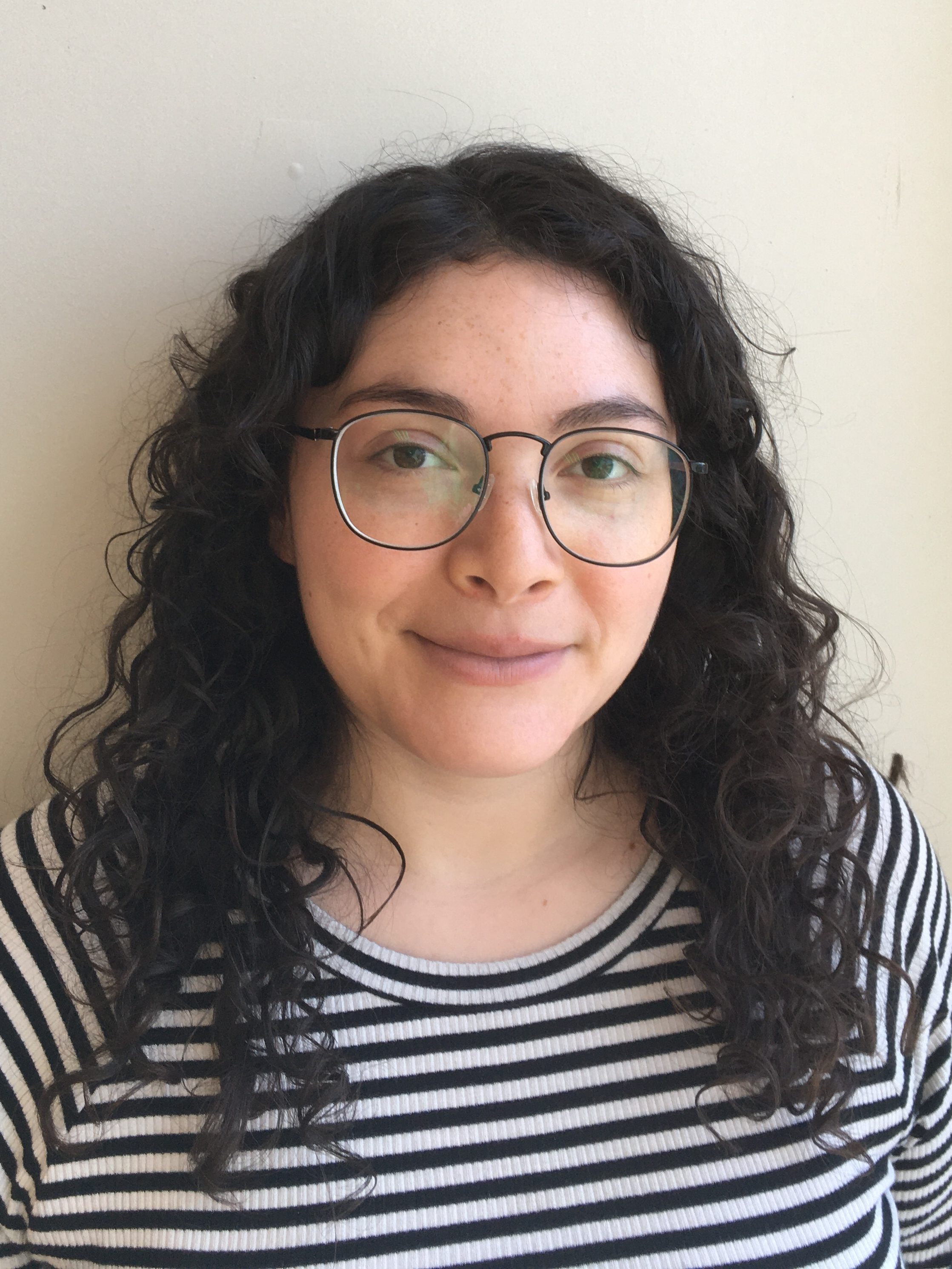 Photograph of Nina, a white-Latinx nonbinary person smiling serenely directly into the camera. Nina has curly dark brown hair that falls to her shoulders, glasses, and is wearing a black and white striped shirt.